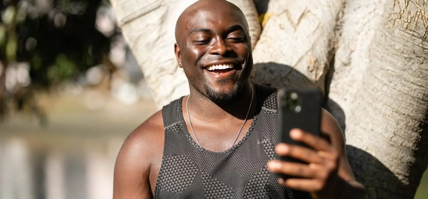 Young black man in a black tank top, under the sun, holding a cell phone and smiling, while receiving TikTok views.
