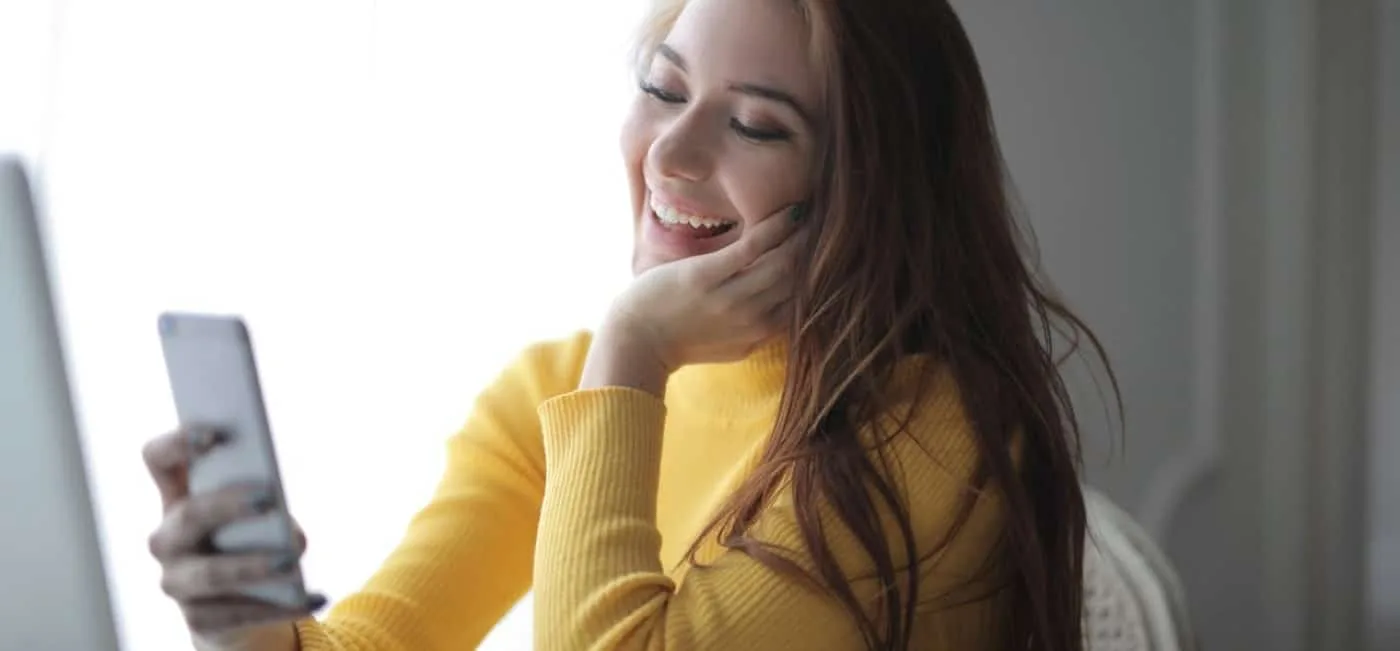 A young woman with long brown hair, wearing a long-sleeved yellow shirt, sits at the living room table, cell phone in hand. She smiles as she learns how to buy TikTok views.