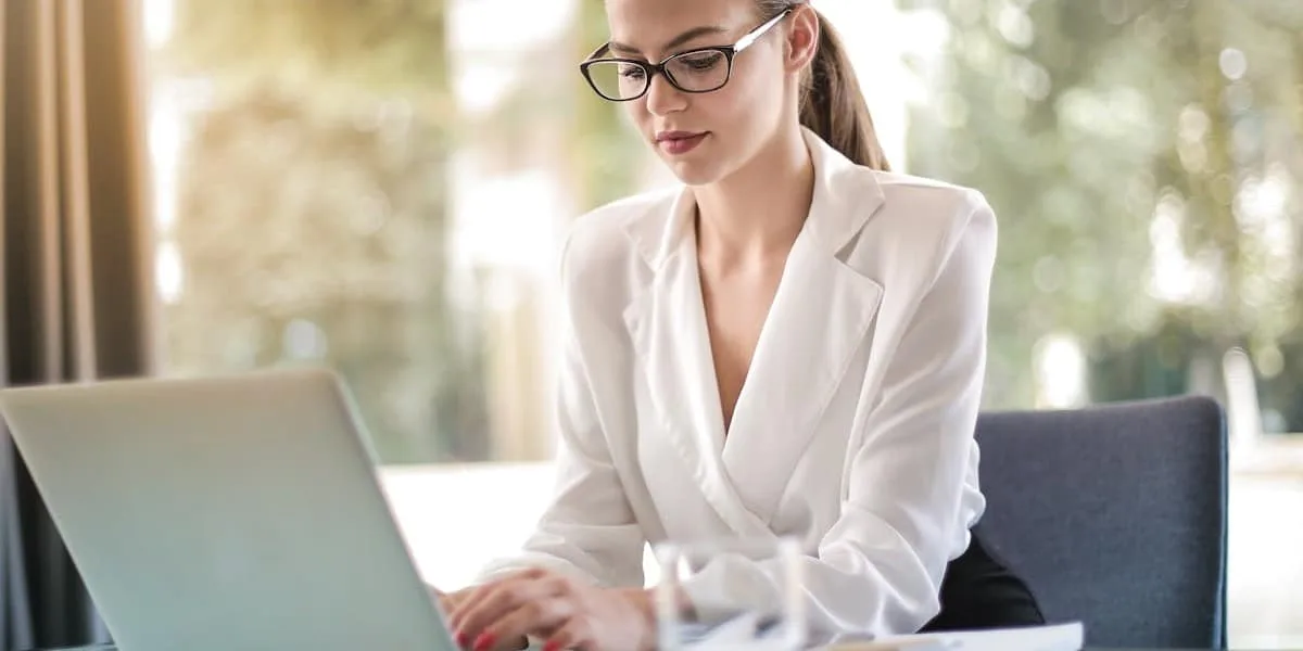 A young entrepreneur wearing a white dress shirt, her hair tied in a ponytail and glasses, is typing on her laptop at her office desk.