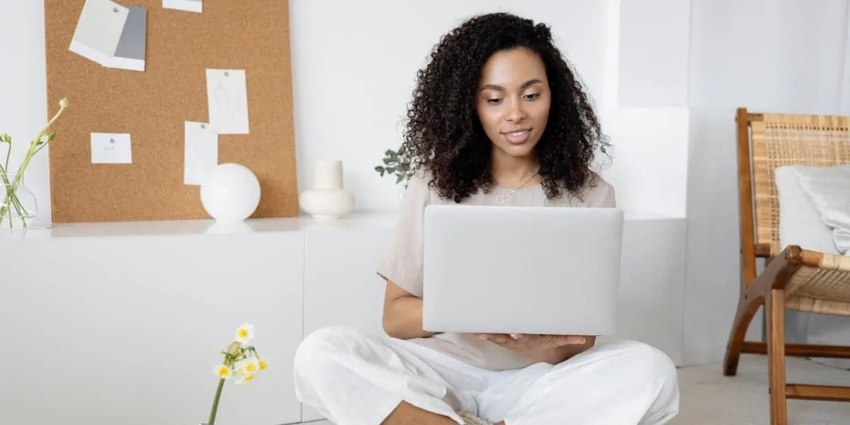 A Black woman dressed in white, sitting cross-legged on the floor, with an open laptop on her lap.