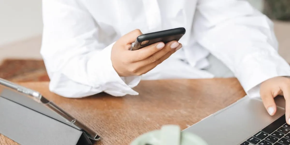 A businesswoman, wearing a white blouse, holds a black cell phone while fiddling with her laptop in the other hand.