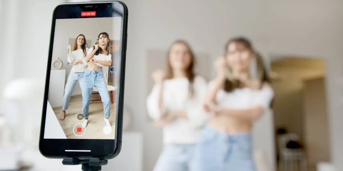 Duas jovens criadores de conteúdo gravando uma correografia para o TikTok. Vemos o celular em primeiro plano, com a imagem de ambas no vídeo, e o fundo está desfocado.