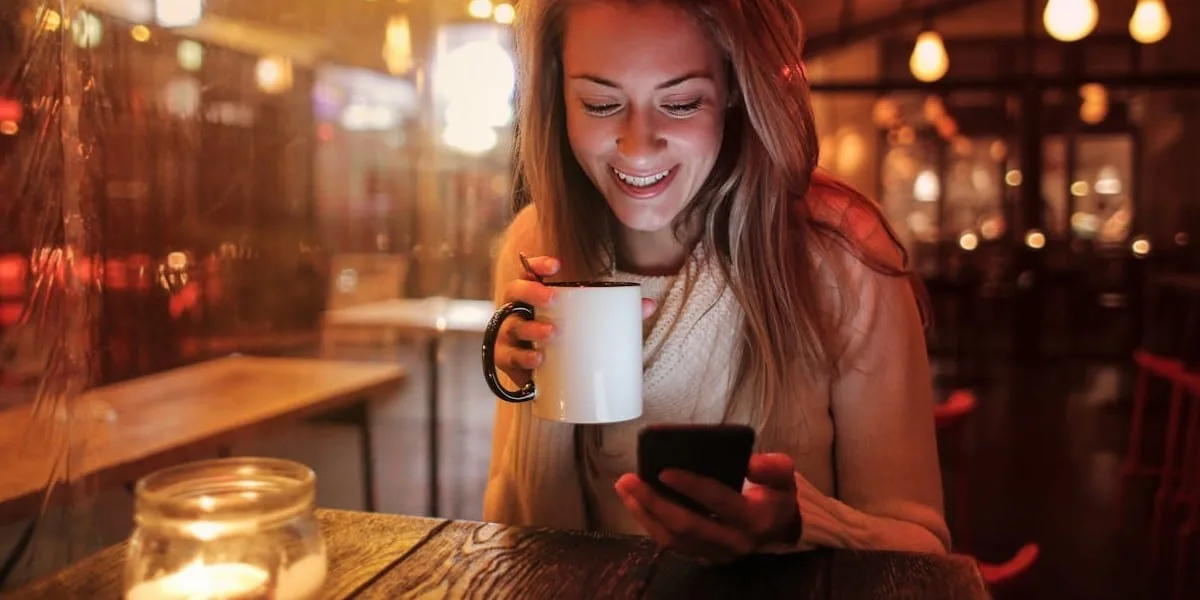 A young blonde woman smiles, holding her cell phone in one hand and a white mug of coffee in the other, in a cozy restaurant. Close-up shot with a blurred background.
