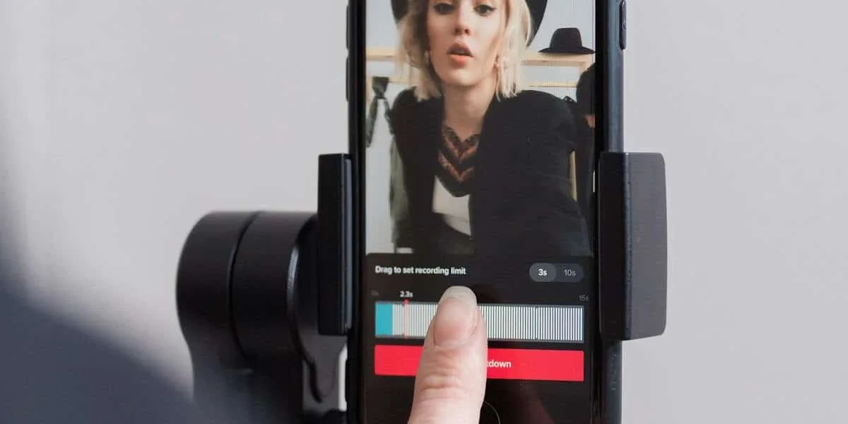 A cell phone is stuck in a holder while a content creator edits a video for Instagram. On the screen, we see a blonde woman in a black hat and blazer, while the user edits some settings on a blue and white bar.