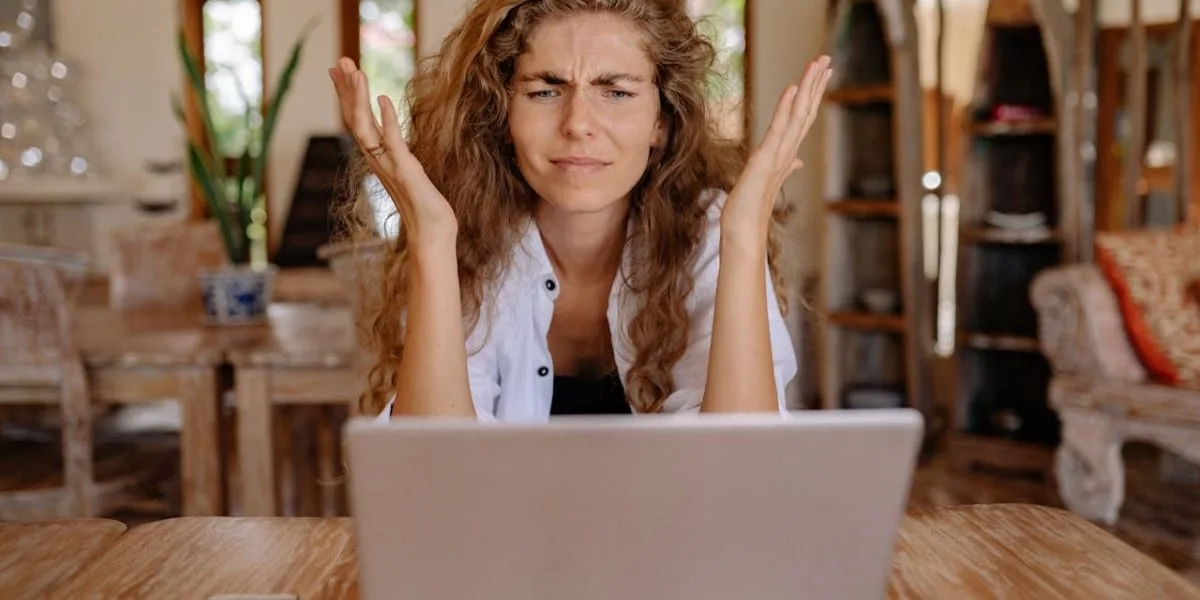 A woman with curly brown hair, sitting in front of her laptop, with her cell phone on the table, and both hands raised, looking confused.