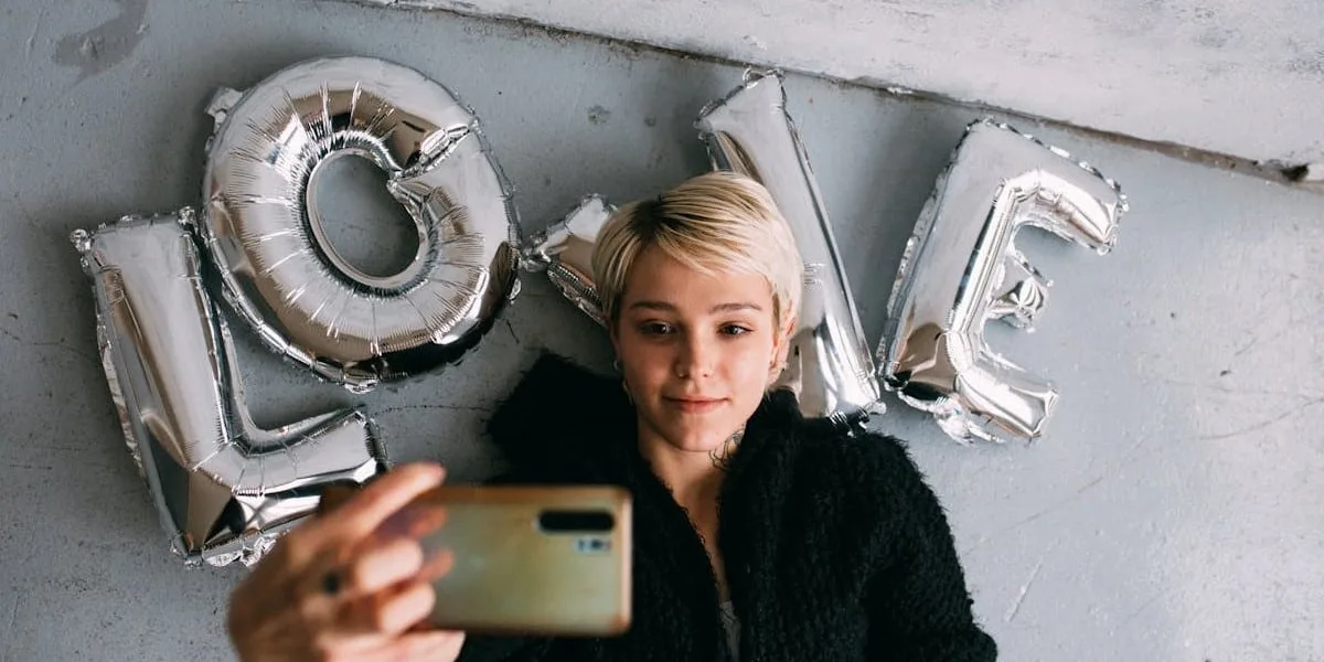 A young blonde woman with short hair, wearing black clothes, lies on silver cushions that spell out the word