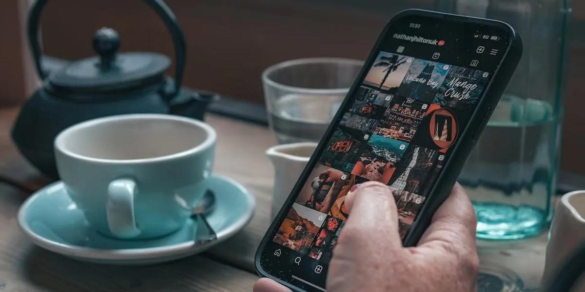 A man is holding his cell phone, with the screen showing his Instagram gallery, choosing an image to post. In the background, we see a coffee table with a cup, a water pitcher, a body, and a kettle.