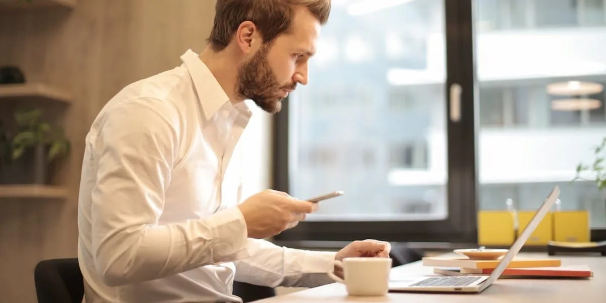 A man in a white dress shirt, seen in profile, is sitting in front of his laptop, holding his cell phone, choosing his Instagram name.