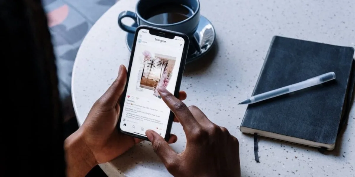 A person is sitting at a coffee table, holding their cell phone and looking at an Instagram post. The scene is viewed from above, behind the person.
