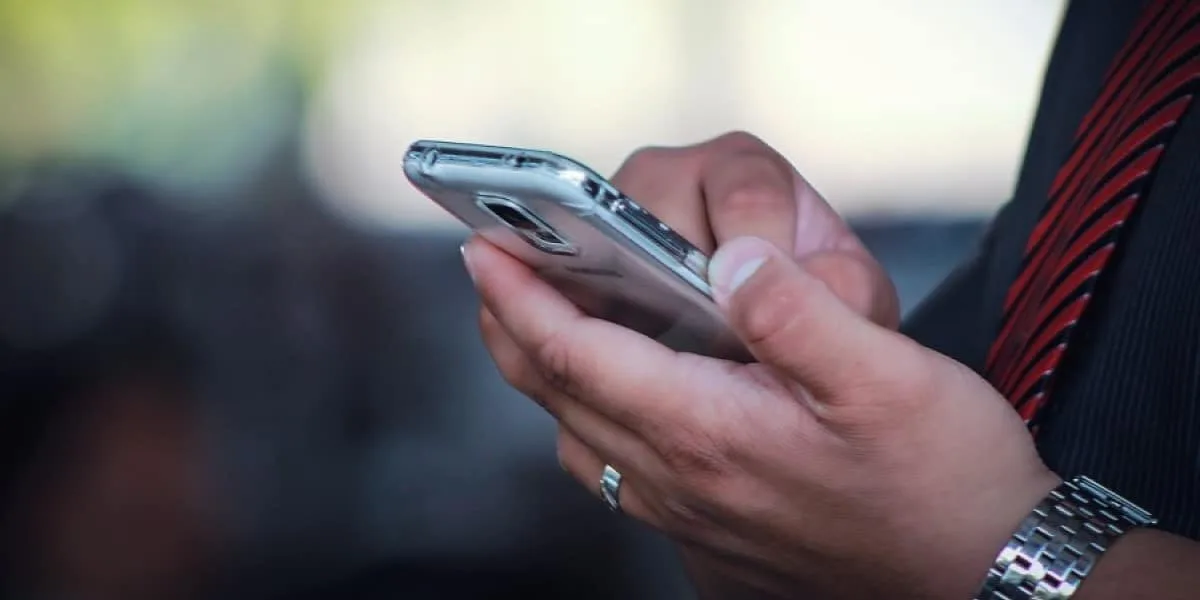 A close-up shot shows a cell phone in the hands of a man wearing a watch and formal attire. His face is not visible. The background is blurred.
