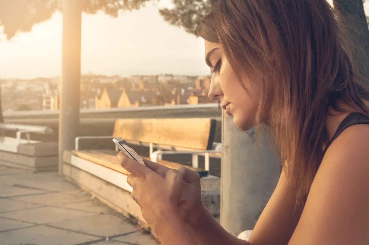 Young blonde woman, seen in profile, using her cell phone on a high balcony. In the background, we see a city landscape.