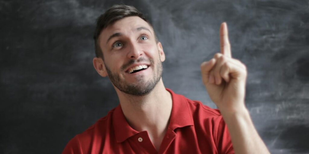Homem jovem usando camisa vermelha, com o braço e o dedo indicador erguido para cima, sorrindo enquanto olha para o alto, com a expressão de quem está tendo uma ótima ideia. Nomes para Instagram masculino.
