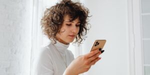 Mulher vista de lado, com cabelos cacheados curtos e blusa branca, enquanto o olha o celular. Ao fundo, vemos paredes brancas. O que é feed no Instagram?