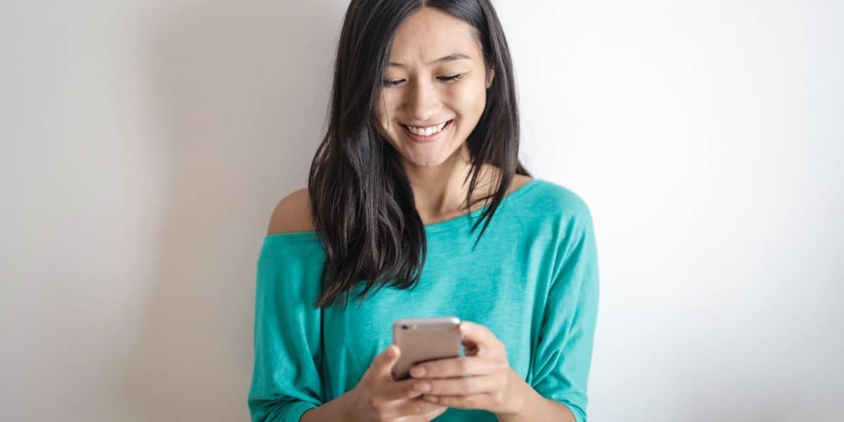 Jovem morena clara, de cabelos lisos médios e pretos, usando blusa verde, mexe no celular para atualizar o Instagram, enquanto sorri. Como atualizar o Instagram.