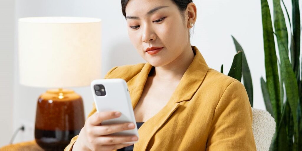 Jovem mulher oriental de blazer amarelo e cabelos curtos, mexendo no celular branco para aprender como tirar o online do Instagram.