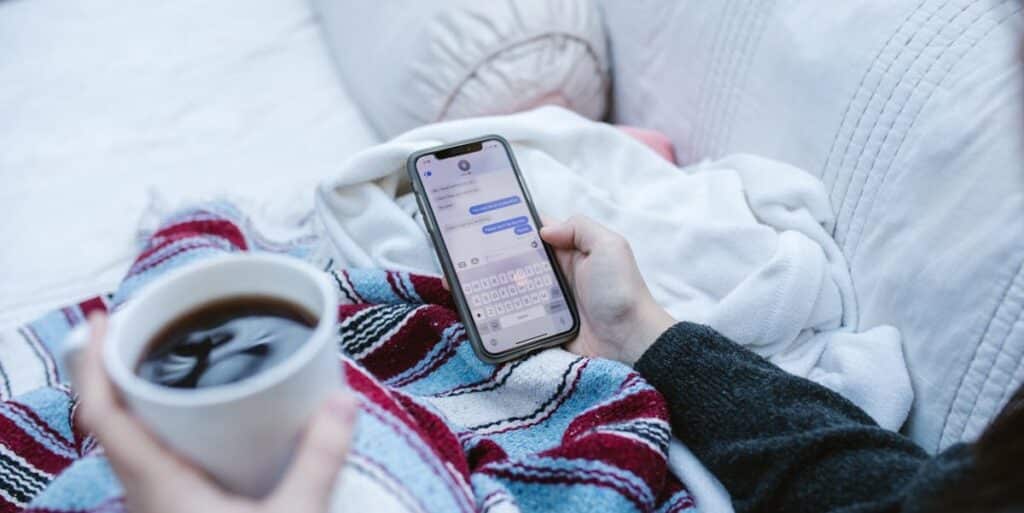 Pessoa enrolada em cobertas sobre a cama, vista por trás, com uma caneca de café e o celular na mão, digitando mensagens no Direct do Instagram. Vemos apenas suas mãos e cobertas. Como apagar conversa do Instagram.