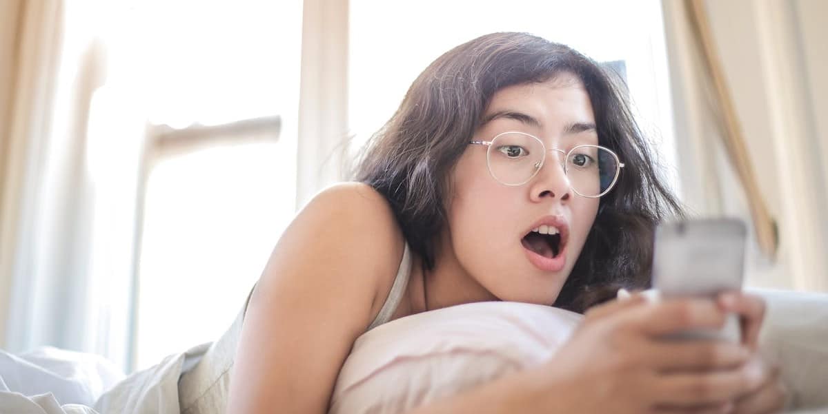 Jovem garota de cabelos castanhos escuros, usando óculos, deitada sobre a cama, coberta com edredom, fazendo uma expressão de grande surpresa, enquanto olha para o celular que segura com as mãos. Como saber se fui bloqueado no Instagram.