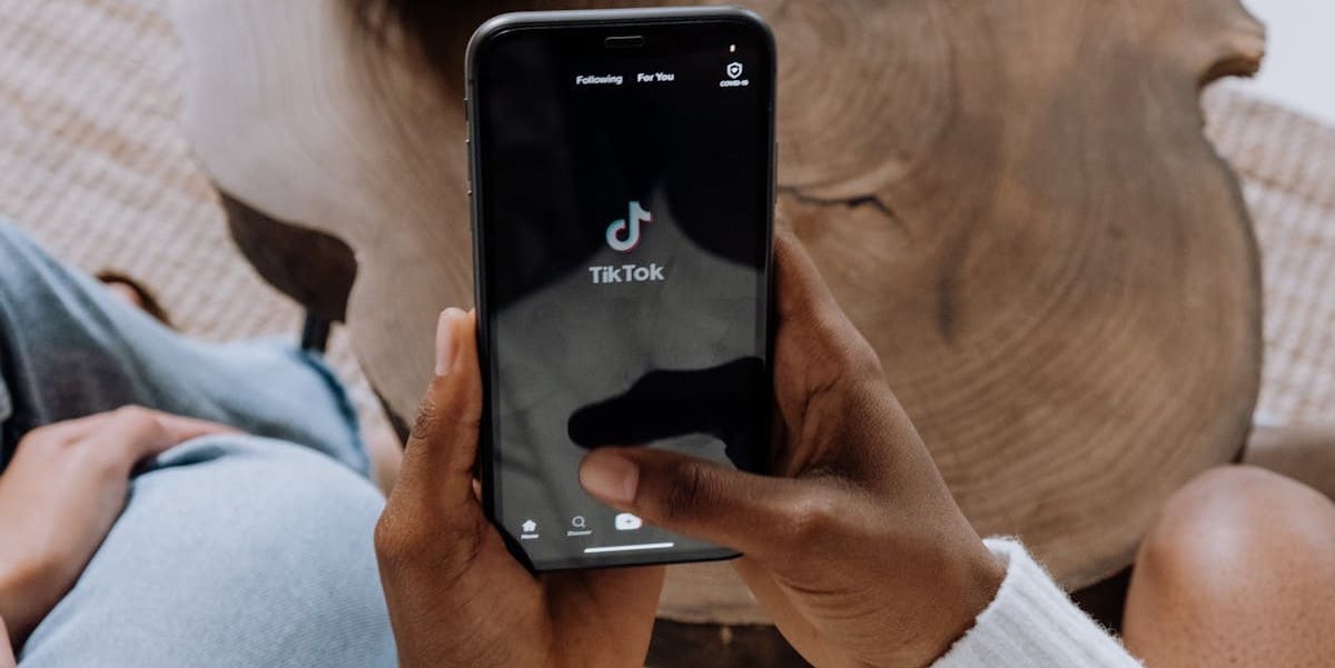 Usuário segurando telefone celular, com tela ligada no app TikTok, que exibe uma imagem preta com o logo do TikTok no centro. Vemos apenas as mãos da pessoa, imagem tirada de ângulo superior. Como baixar vídeo TikTok.