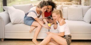Três jovens amigas em sala de estar, duas sentadas no sofá e uma chão, enquanto uma delas mostra uma publicação no Instagram no celular para as outras. Todas estão empolgadas. Como repostar no Instagram.