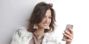 Jovem mulher bem arrumada, segurando seu telefone, sorrindo enquanto faz downloads do Instagram, com um dos aplicativos para baixar Status do Instagram.
