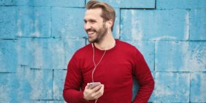 Homem de roupa vermelha, encostado em parede azul na rua, ouvindo música com fones de ouvido no celular. Como colocar musica na bio do Instagram