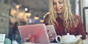 Jovem loira, com blusa vermelha, sorri enquanto segura o celular, em uma cafeteria, com o computador logo a frente e uma xícara de café. Como funciona a ordem de visualização dos stories.