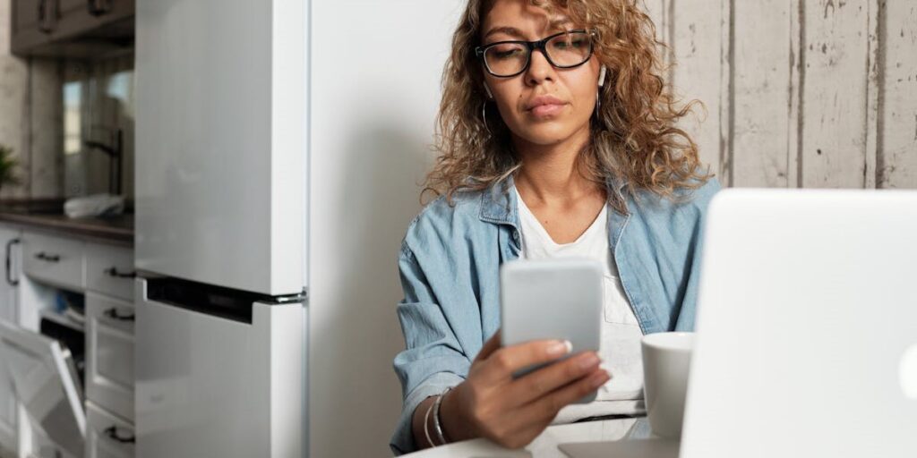 Mulher morena de cabelos castanhos, usando óculos e roupas casuais, segura um celular branco para conferir seu Instagram stories. Ela está sentada na mesa da sala de jantar com seu notebook branco logo a frente. Como saber se fui bloqueado no Instagram Stories.