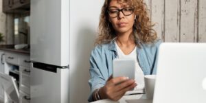 Mulher morena de cabelos castanhos, usando óculos e roupas casuais, segura um celular branco para conferir seu Instagram stories. Ela está sentada na mesa da sala de jantar com seu notebook branco logo a frente. Como saber se fui bloqueado no Instagram Stories.
