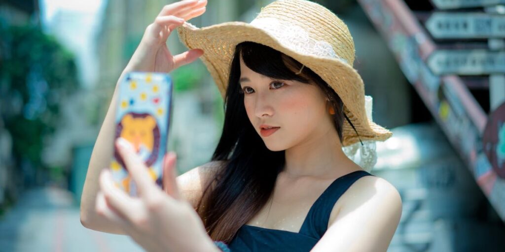 Jovem de pele clara e cabelos escuros, usando chapéu de palha, tira selfie em ambiente urbano ensolarado. Como trocar a foto do perfil do Instagram.