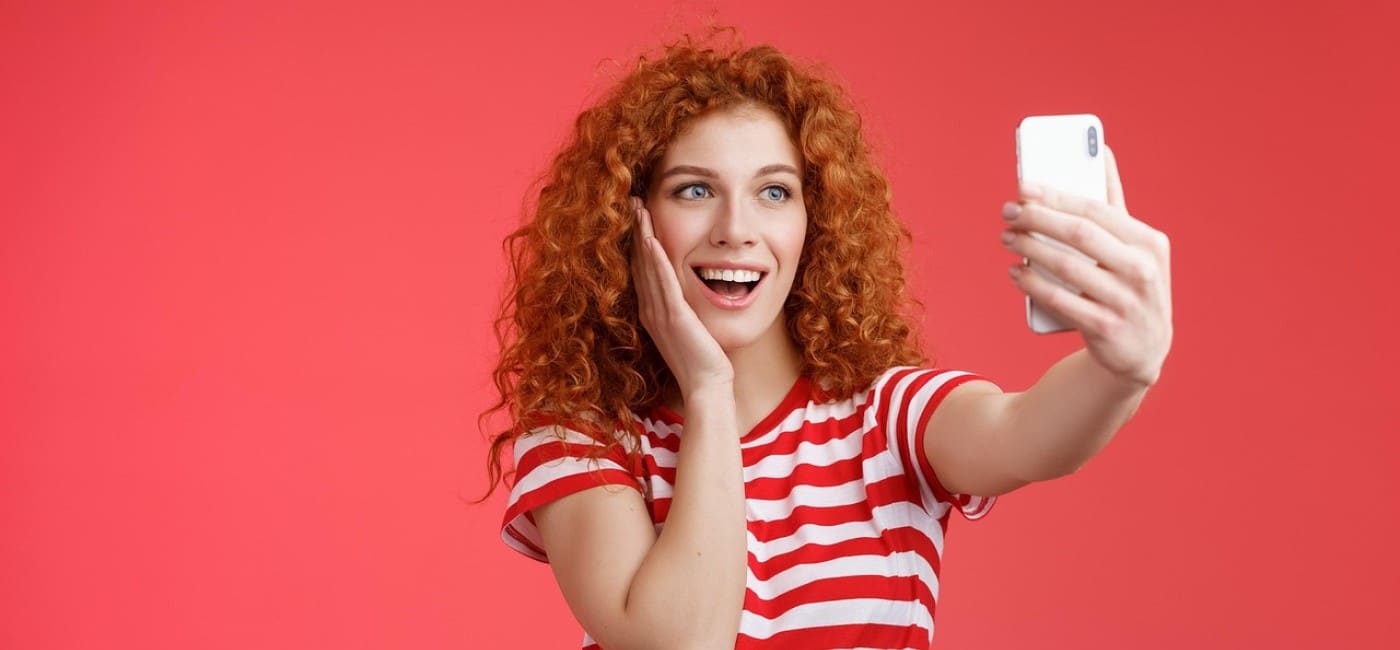 Moça ruiva de cabelo cacheado, com blusa de listras vermelhas e brancas, segura celular na altura dos olhos, sorrindo, admirada, enquanto recebe visualizações reels Instagram