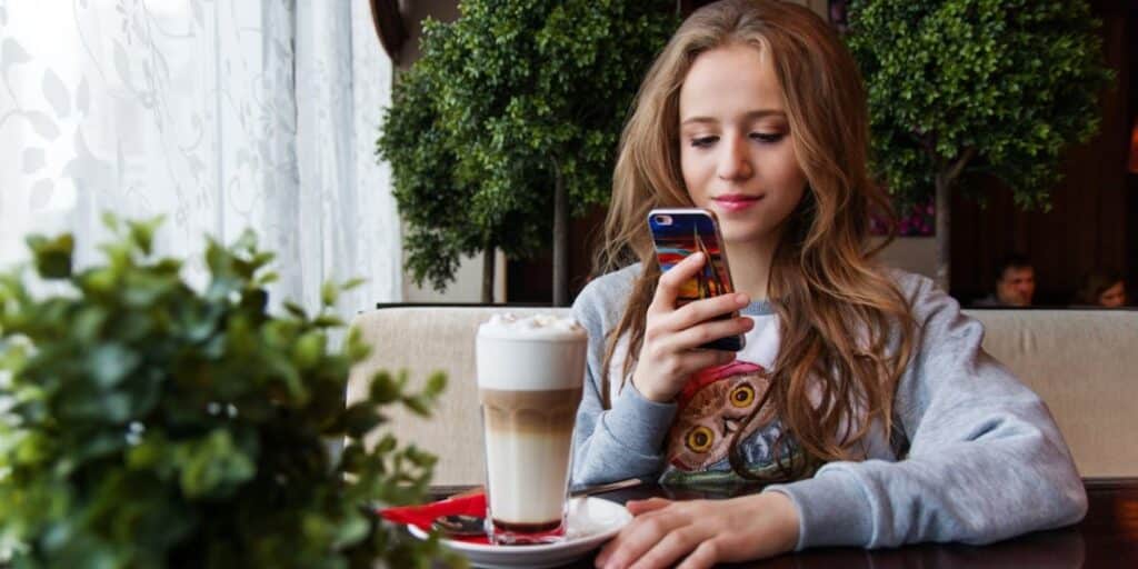 Jovem de cabelos claros e roupas casuais, tomando um café gelado em uma cafeteria, enquanto confere o Instagram no celular. Como saber se uma pessoa está online no Instagram