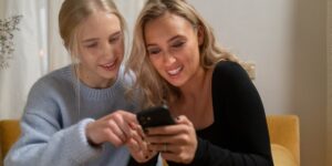 Duas jovens loiras sorrindo, usando roupas casuais, empolgadas, olhando as visualizações da conta de uma delas no celular. O que é impressões do Instagram.