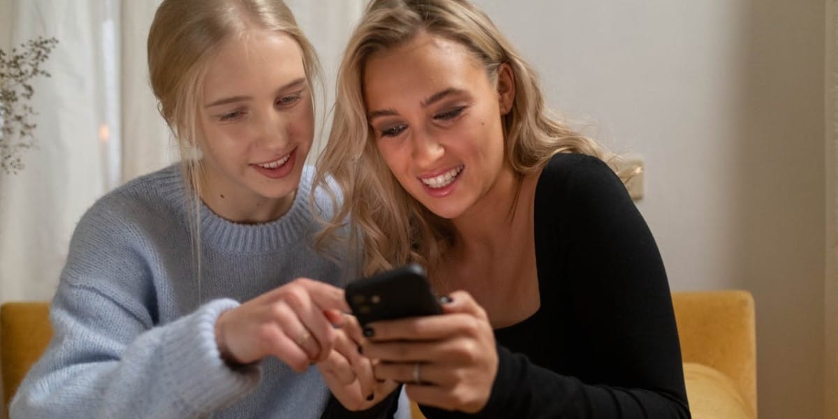Duas jovens loiras sorrindo, usando roupas casuais, empolgadas, olhando as visualizações da conta de uma delas no celular. O que é impressões do Instagram.