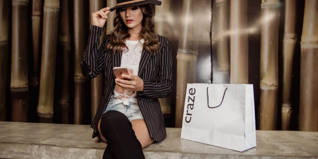 Jovem com estilo moderno, usando chapéu, ao lado de uma sacola de compras, sentada enquanto olha o celular. O que significa painel profissional no Instagram