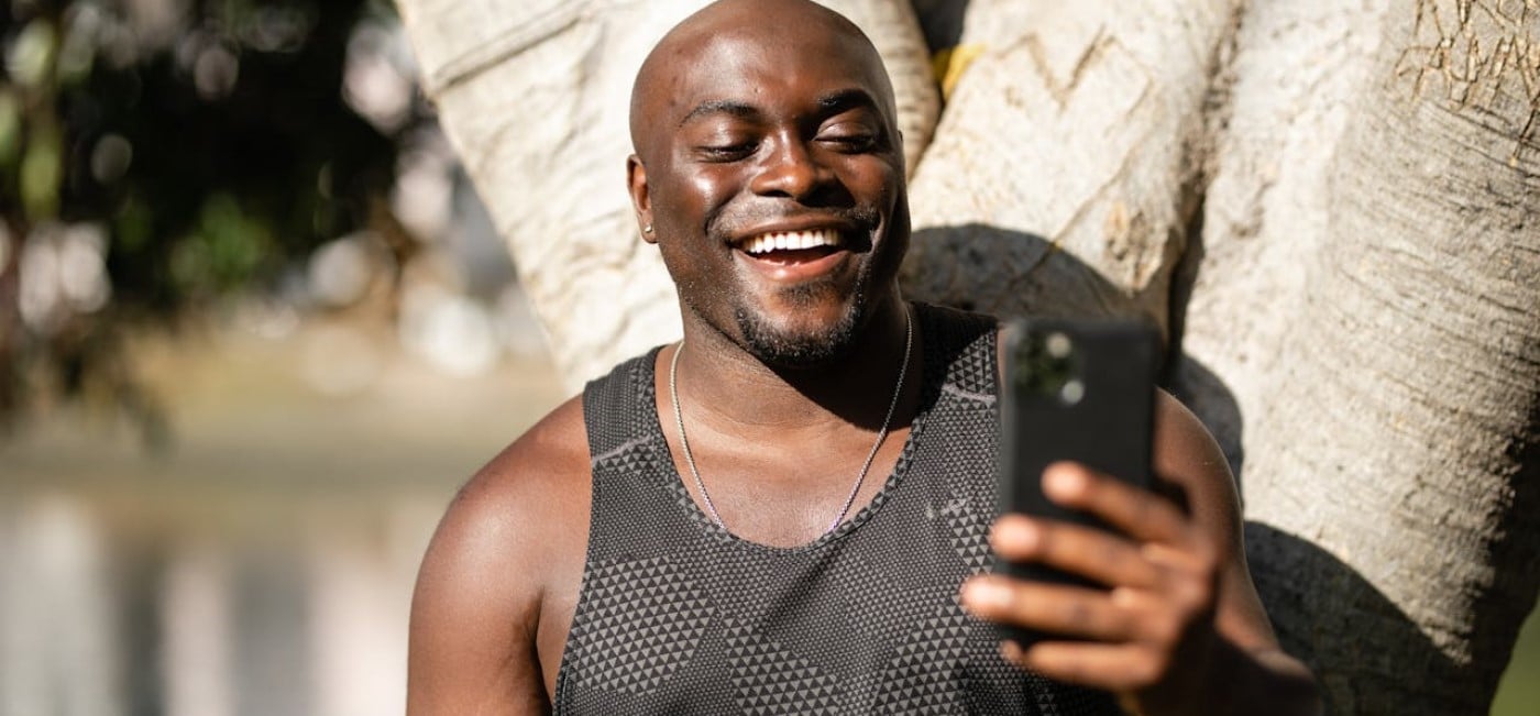 Jovem homem negro de camisa regata preta, sob o sol, segurando celular e sorrindo, enquanto recebe visualizações TikTok
