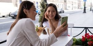 Jovem com celular sorri e mostra os seguidores comprados chegando em seu perfil para uma amiga em café